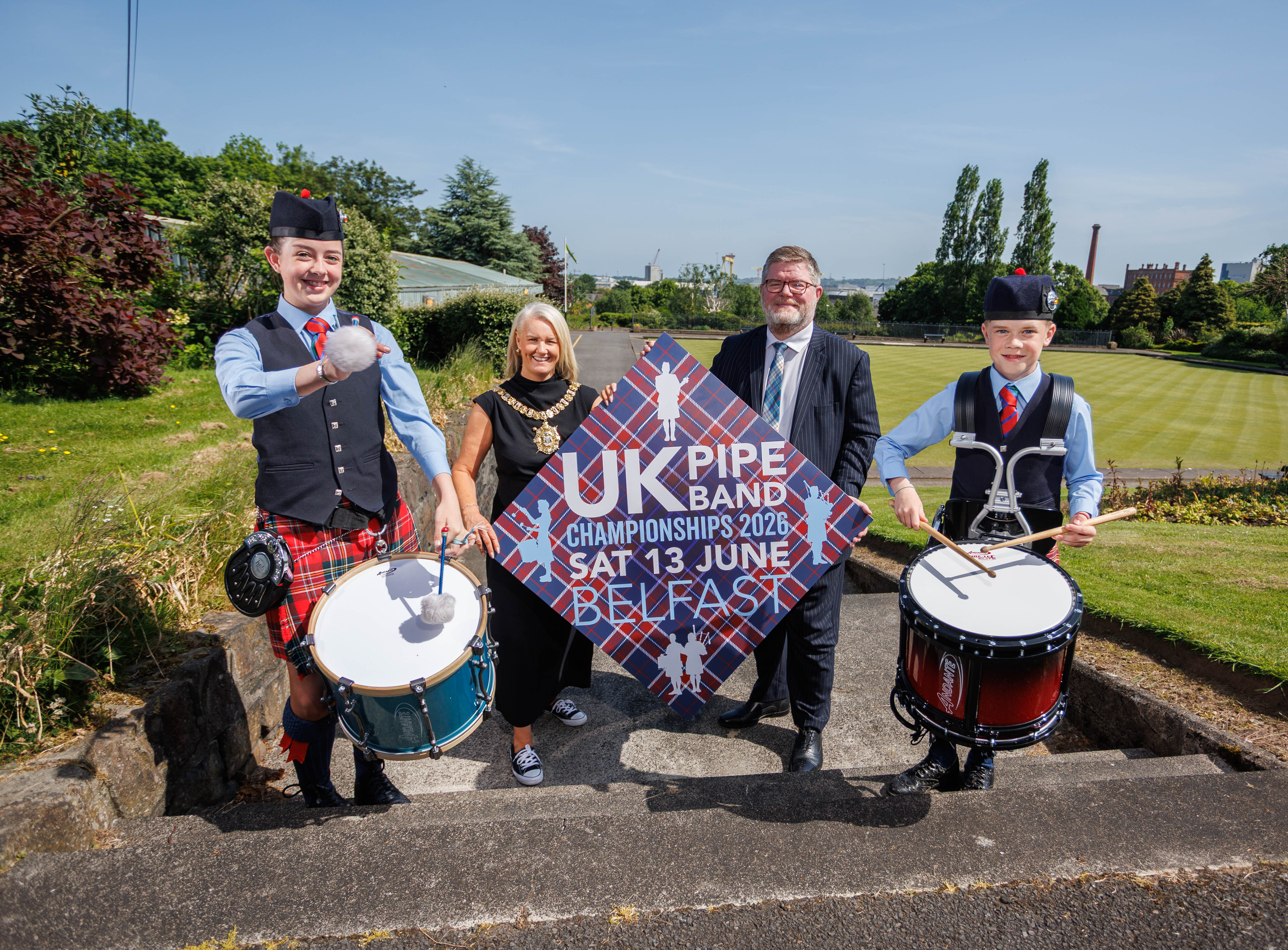 Lord Mayor of Belfast wearing a black dress and her ceremonial chain stands beside a man in a black suit. They are holding a large diamond-shaped sign for the UK Pipe Band Championships 2026 in Belfast on Saturday 13 June. Two drummers in traditional Scottish attire stand either side.
