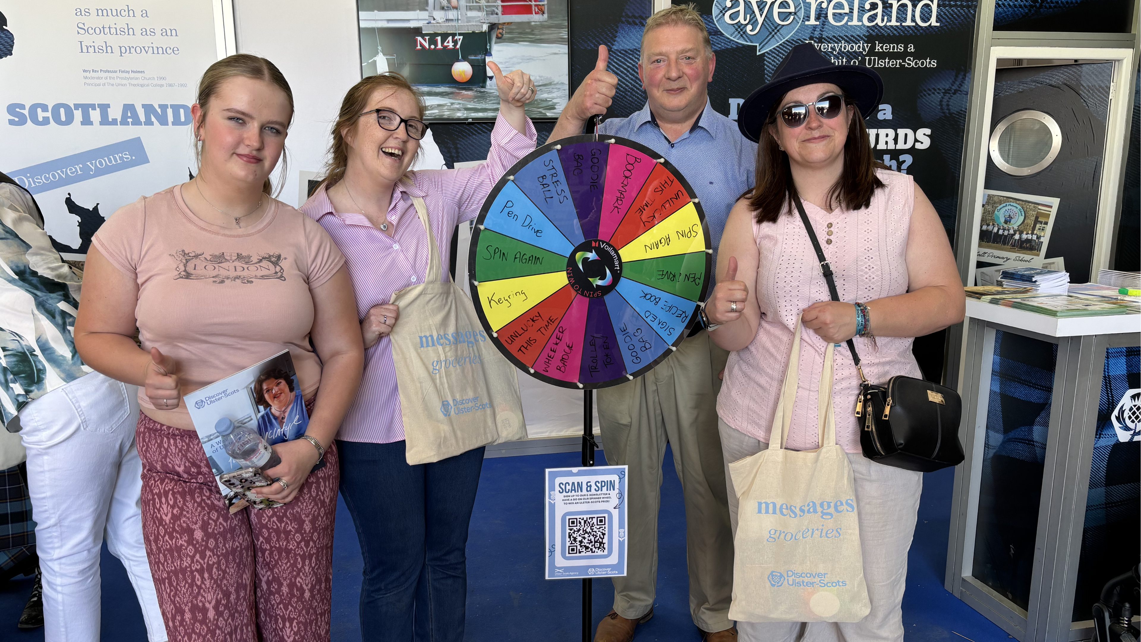 Three women and a man standing indoors around a colourful prize wheel. Each is giving a thumb-up gesture and holding cream tote bags which say 'Messages' in light blue text.