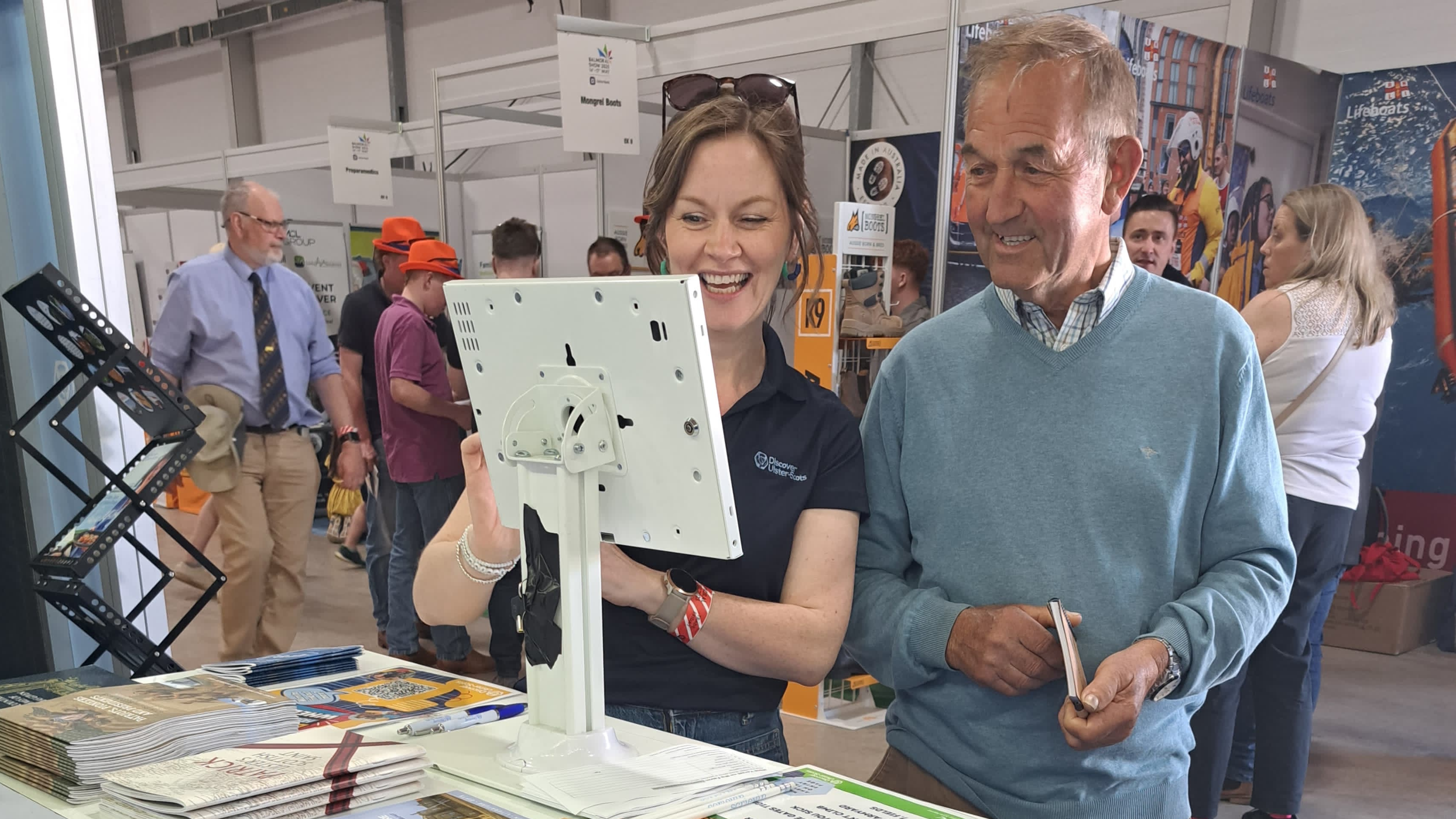 A man and a woman interact with a touchscreen display at a busy indoor exhibition booth.