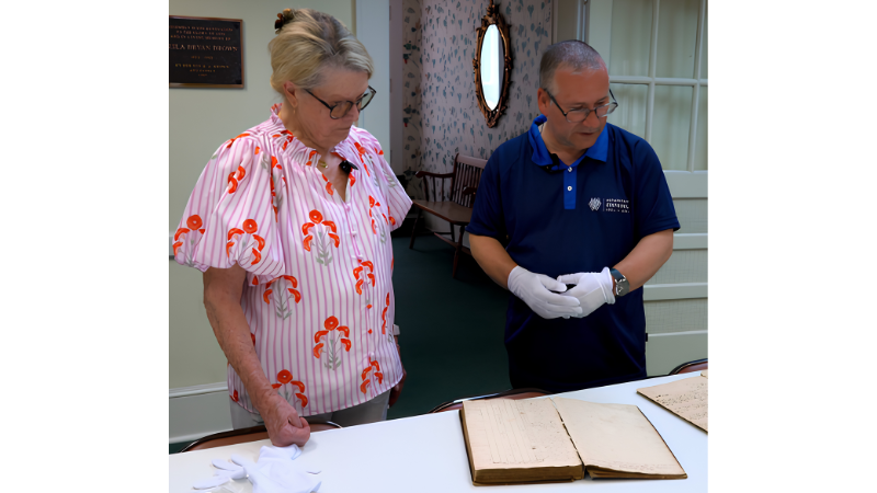 A man and a woman examine old, open books on a table in a room with vintage decor and floral wallpaper. The woman, left, wears a pink and white striped blouse. The man, right, is wearing a navy polo t-shirt.
