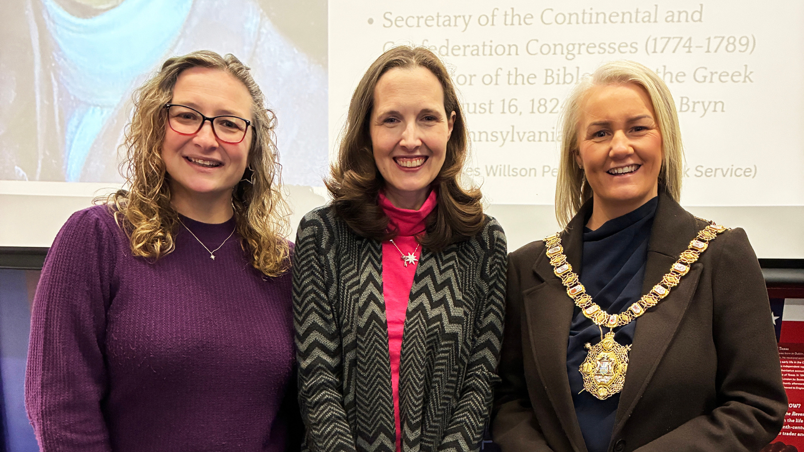 Three women standing indoors in front of a presentation screen. The woman on the right is wearing a ceremonial chain of office around her neck.