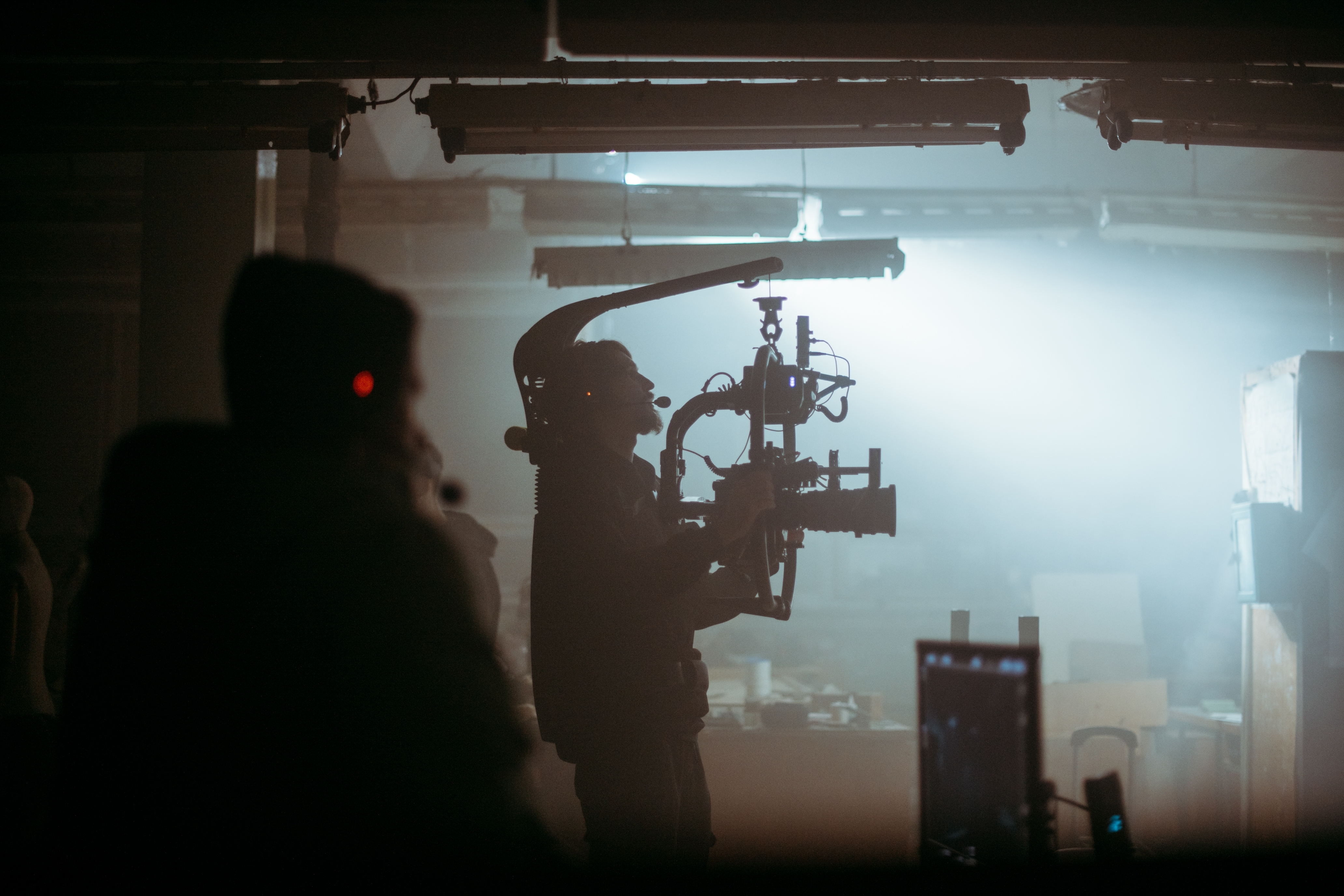 Silhouetted camera operator filming with a stabiliser rig in a dimly lit, foggy indoor set.