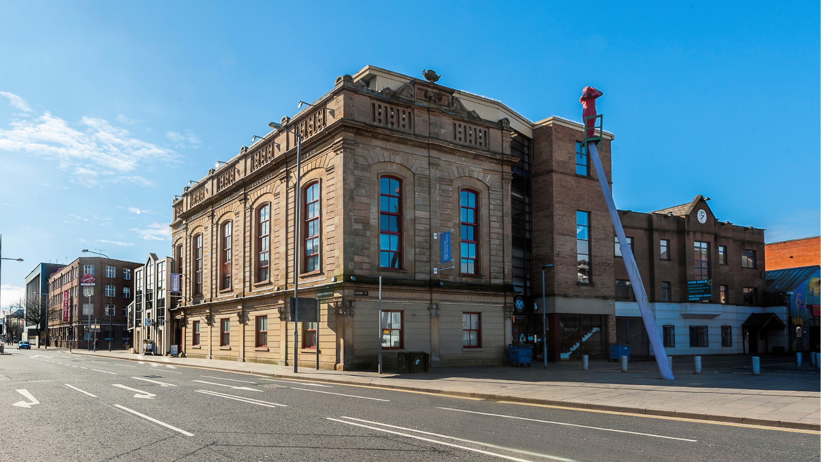 Historic stone building, The Corn Exchange in Belfast, with large windows under a clear blue sky on a quiet street.