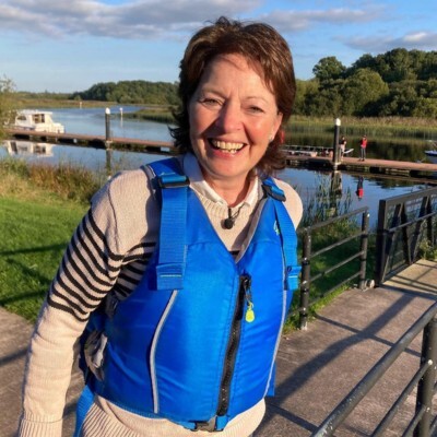 A woman wearing a bright blue life jacket and standing near a calm river with boats and green trees in the background under a partly cloudy sky.