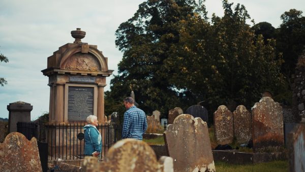 A man and woman standing near a large historic monument surrounded by old gravestones in a cemetery. There are a number of tall trees in the background.