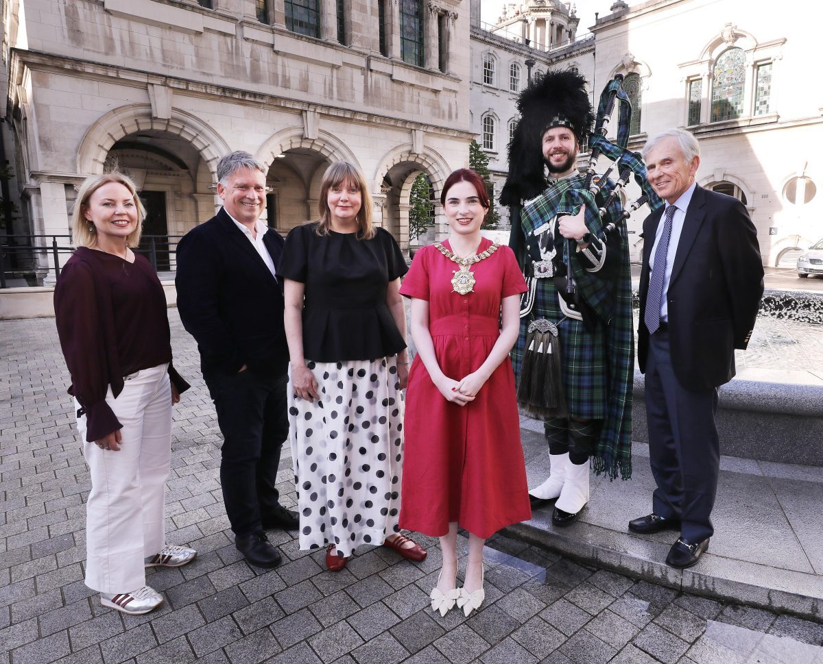 Group of six adults standing outdoors in front of Belfast City Hall, a historic stone building, including a man dressed in traditional Scottish attire playing bagpipes.