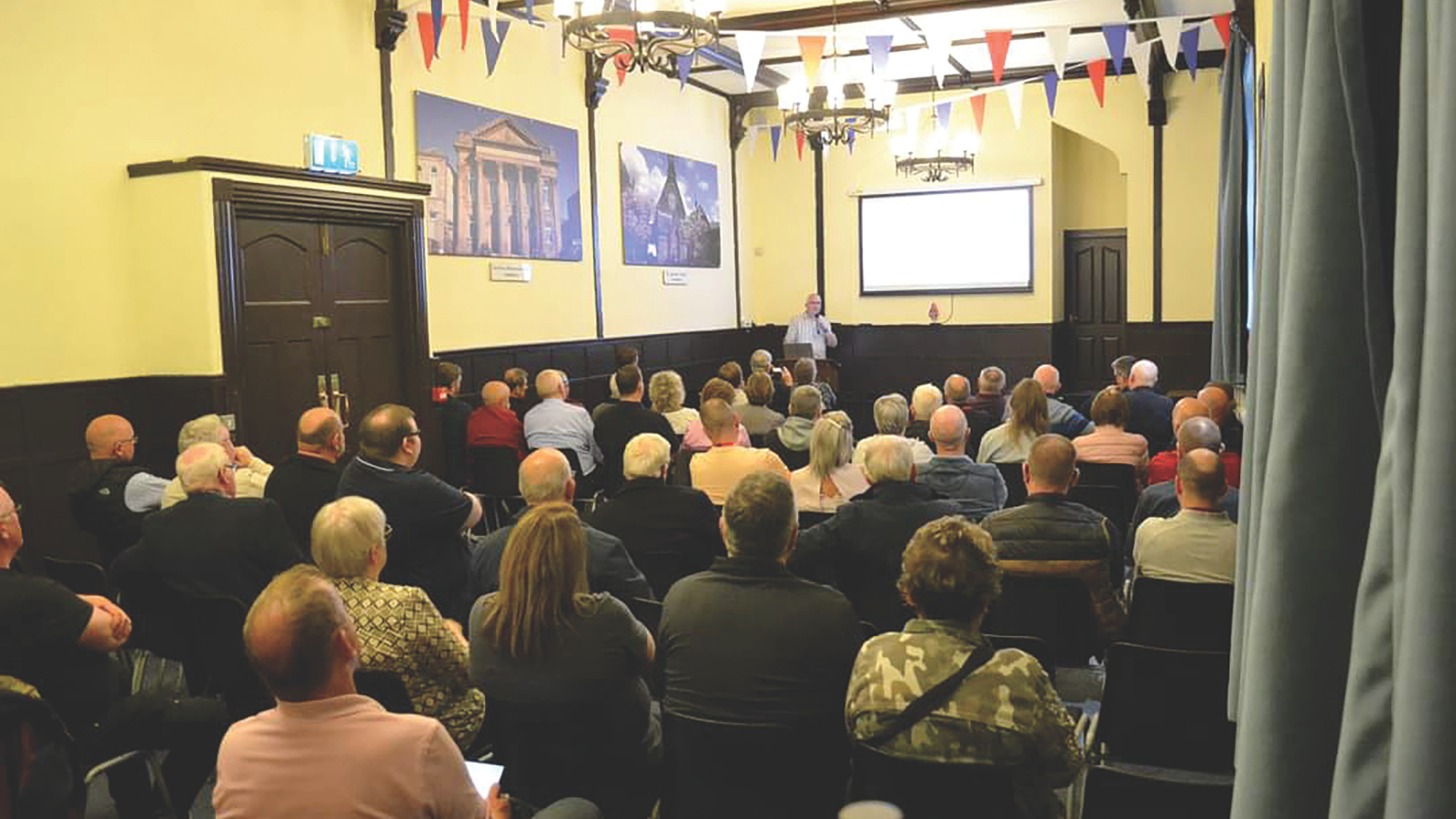 Audience attentively listening to a speaker at a historical talk. The interior walls of the building are decorated with historical photos of other buildings.