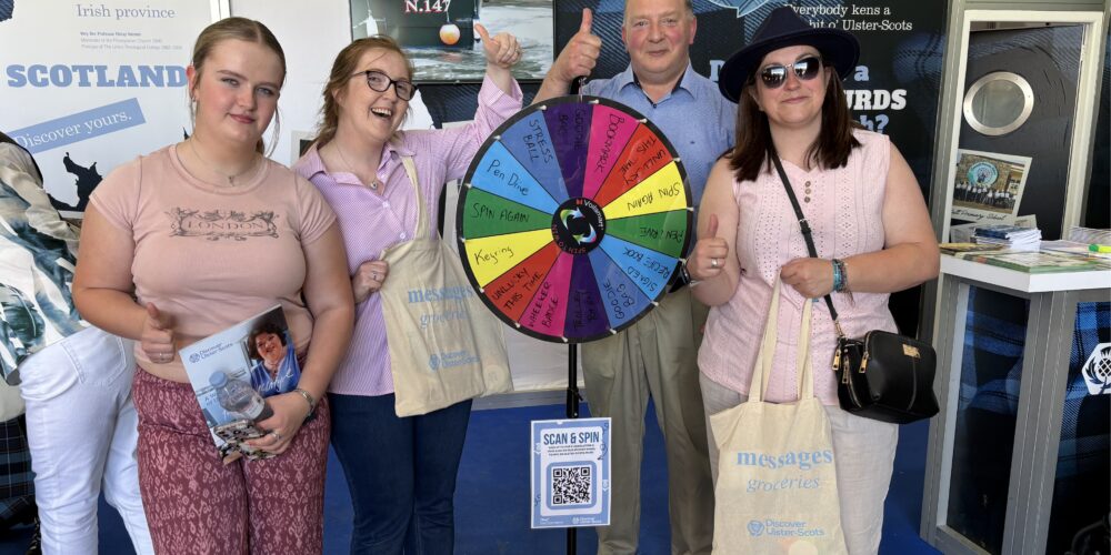 Three women and a man standing indoors around a colourful prize wheel. Each is giving a thumb-up gesture and holding cream tote bags which say 'Messages' in light blue text.
