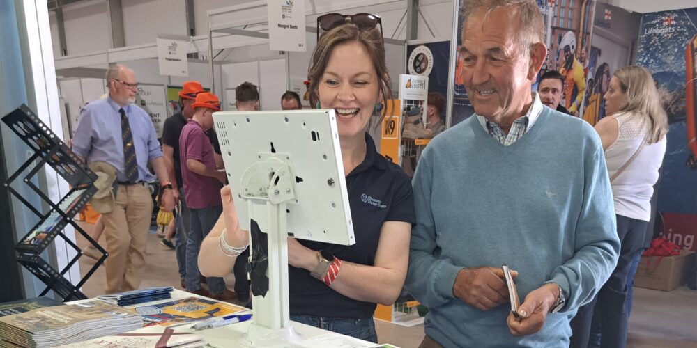 A man and a woman interact with a touchscreen display at a busy indoor exhibition booth.