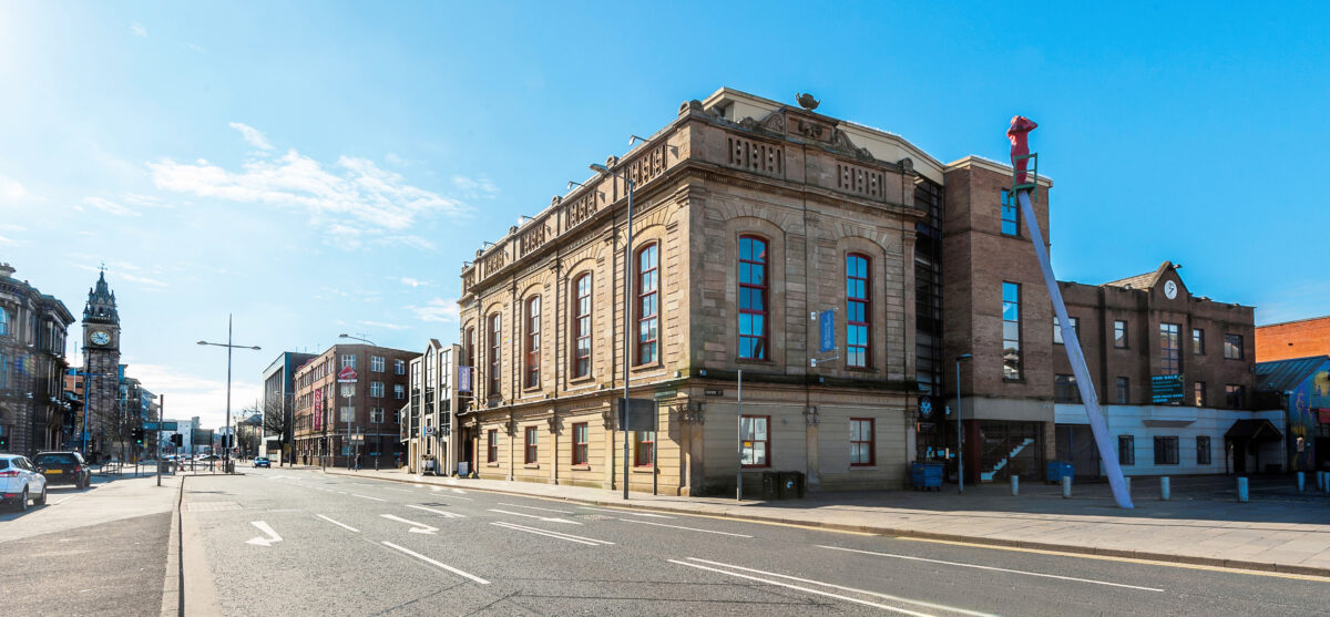 The Corn Exchange Belfast, a historic stone building with large arched windows. It is on a quiet city street under a clear blue sky.