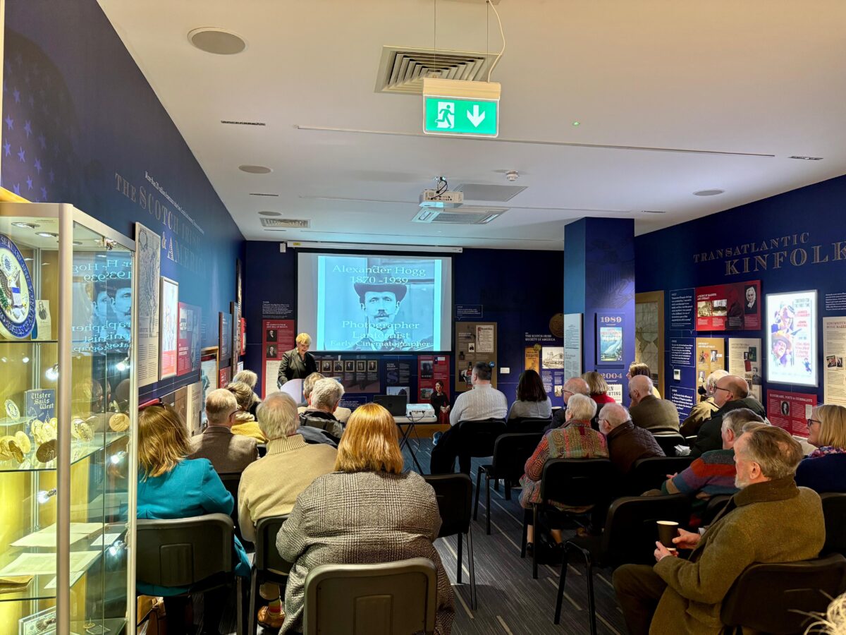 Audience seated in a rows of six in a museum gallery. They are watching a presentation on a historical figure, Alexander Hogg, with information projected on a large wall-mounted screen.