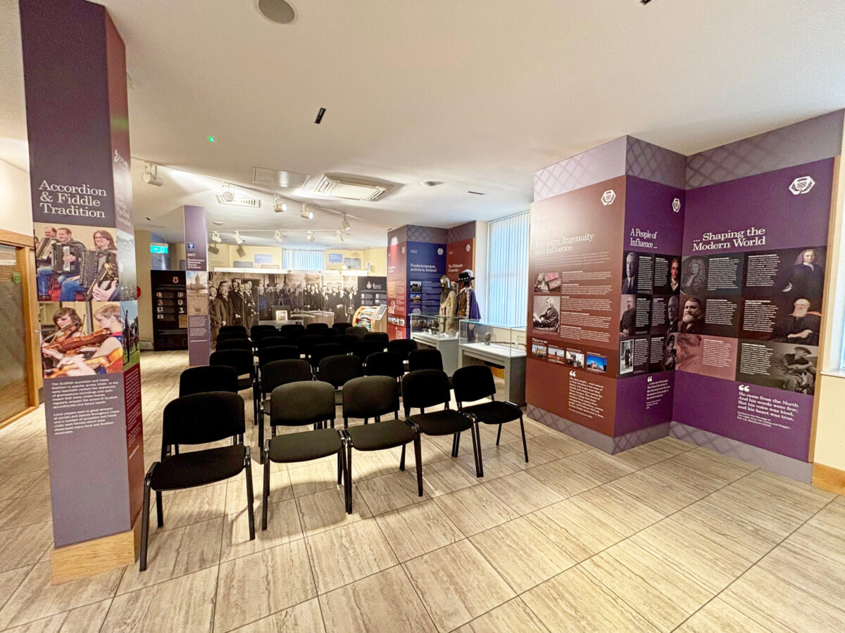 Empty chairs arranged in rows facing an exhibition area with informational panels and historical displays in a museum setting.