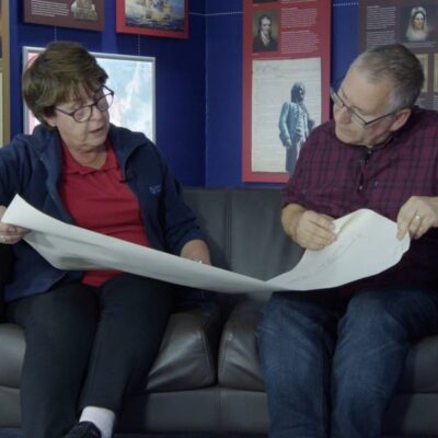 A man and woman seated on a black couch examining a large unfolded map in a room with historical displays on the walls.