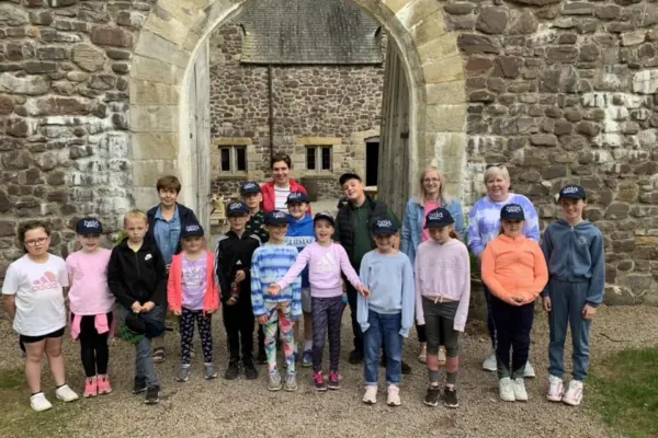 Group of children and two adults standing under a large stone archway in front of a historic brick building on a gravel path. The children are wearing navy baseball caps with 'heid' written on them in white.