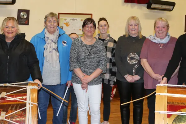 Group of eight women standing indoors a line. They are behind two large Lambeg drums with sticks in hand, ready to play. Photo credit: McAuley Multimedia.