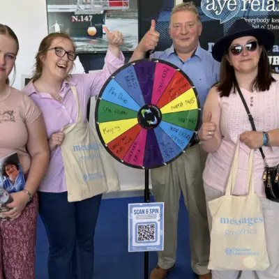 Three women and a man standing indoors around a colourful prize wheel. Each is giving a thumb-up gesture and holding cream tote bags which say 'Messages' in light blue text.