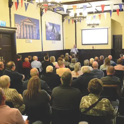 Audience attentively listening to a speaker at a historical talk. The interior walls of the building are decorated with historical photos of other buildings.