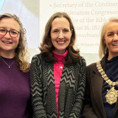 Three women standing indoors in front of a presentation screen. The woman on the right is wearing a ceremonial chain of office around her neck.