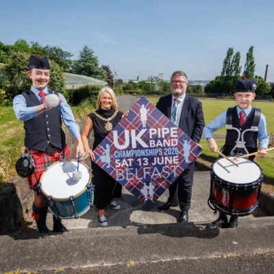 Lord Mayor of Belfast wearing a black dress and her ceremonial chain stands beside a man in a black suit. They are holding a large diamond-shaped sign for the UK Pipe Band Championships 2026 in Belfast on Saturday 13 June. Two drummers in traditional Scottish attire stand either side.