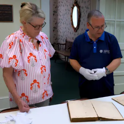 A man and a woman examine old, open books on a table in a room with vintage decor and floral wallpaper. The woman, left, wears a pink and white striped blouse. The man, right, is wearing a navy polo t-shirt.
