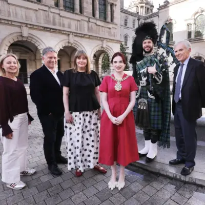 Group of six adults standing outdoors in front of Belfast City Hall, a historic stone building, including a man dressed in traditional Scottish attire playing bagpipes.