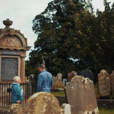 A man and woman standing near a large historic monument surrounded by old gravestones in a cemetery. There are a number of tall trees in the background.