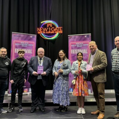 Five people stand on stage at Newgate Arts & Culture Centre holding festival brochures under a neon sign.
