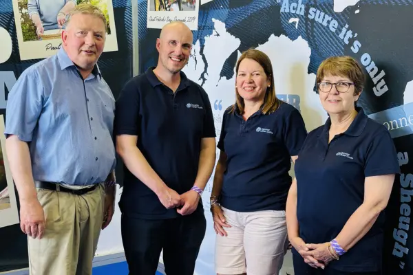 Four adults standing indoors smiling. Three of them wear matching navy blue polo shirts with logos and jeans, and one is in a light blue button-up shirt and beige pants.