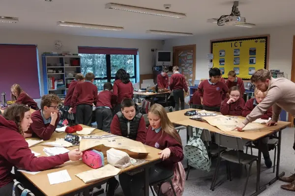 Students in maroon uniforms collaborate on projects at classroom tables while a teacher assists a group nearby.