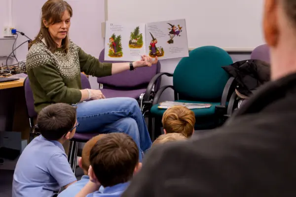 A female teacher in a green sweater reads a colourful storybook to a group of attentive schoolchildren seated on the floor in a classroom.