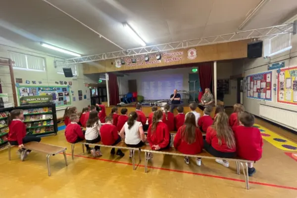 Primary school students in red uniforms sit on two benches facing a stage in a school assembly hall. Two adults stand at the front of the group talking.