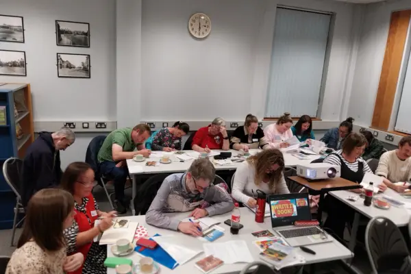 A group of adults seated at tables in a classroom. They are taking part in a workshop and focused on writing and reading materials.