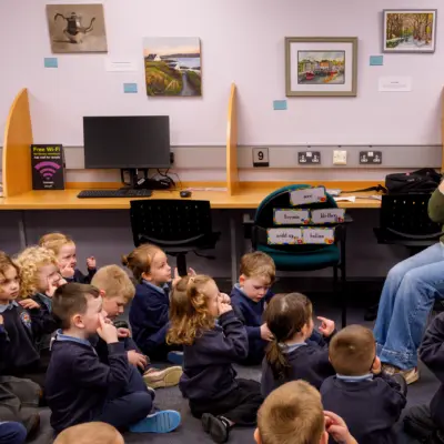 A teacher sits on a chair leading a group of young students. The students are seated on the floor in a classroom.