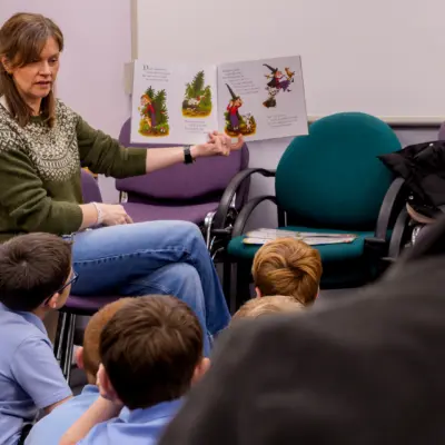 A female teacher in a green sweater reads a colourful storybook to a group of attentive schoolchildren seated on the floor in a classroom.