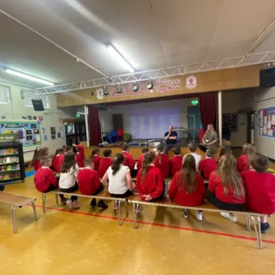 Primary school students in red uniforms sit on two benches facing a stage in a school assembly hall. Two adults stand at the front of the group talking.
