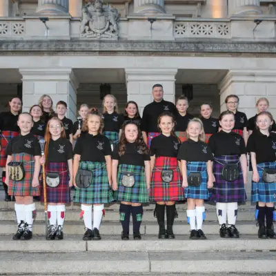 A group of children and adults wearing traditional Scottish kilts in green, blue and red tartans, standing on stone steps in front of Stormont.