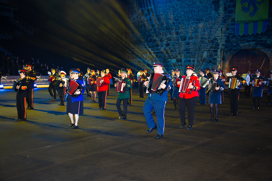 Group of uniformed musicians playing accordions in a night-time outdoor performance. They are standing in formation in 5 rows and in front of a large grey castle backdrop.