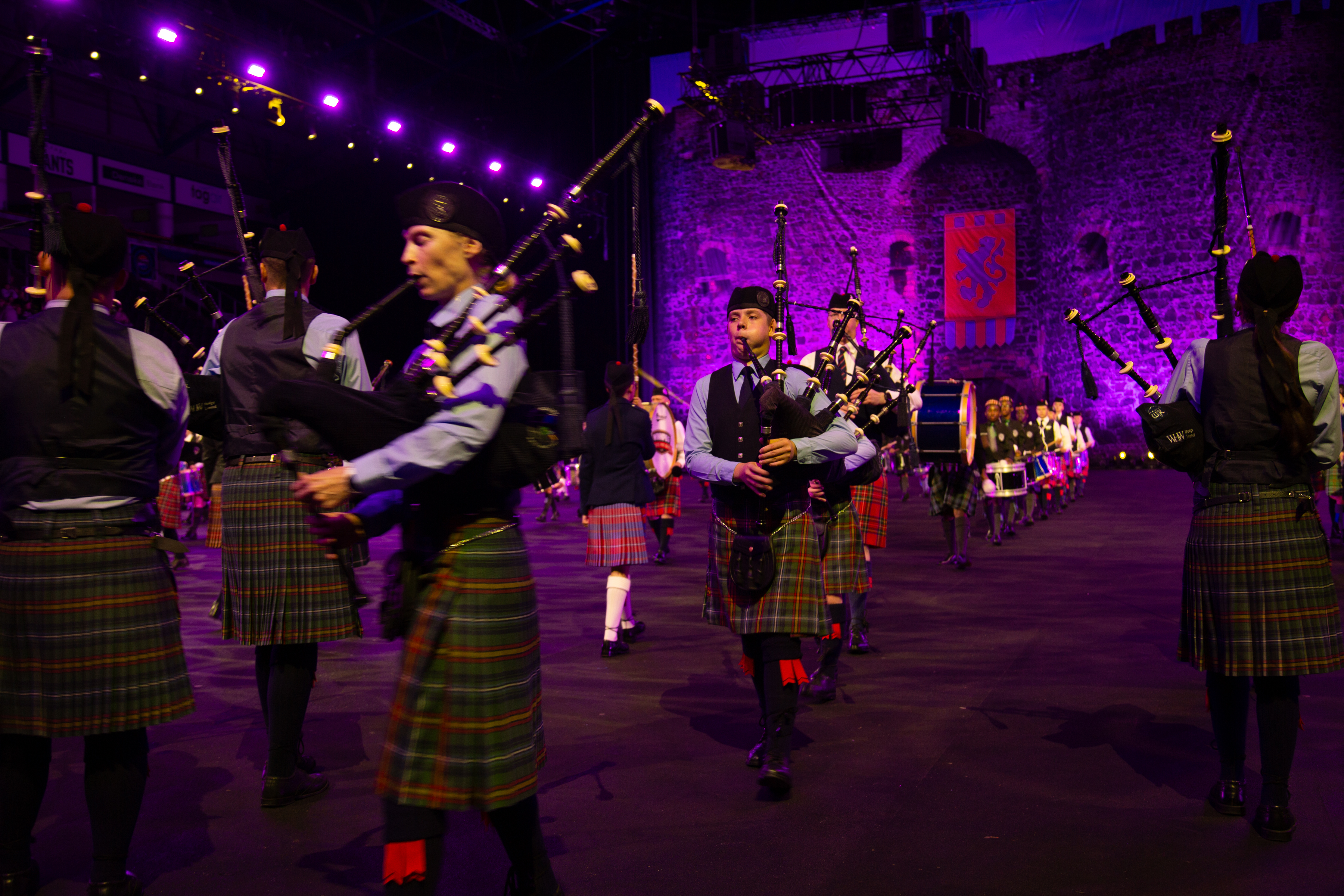 A large group of bagpipers in traditional Scottish kilts and attire stand in various rows, performing under purple stage lights at an outdoor event with a castle backdrop.