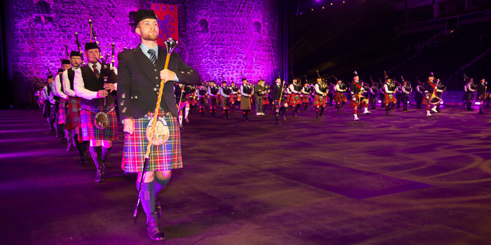 A large group of bagpipers and drum majors in traditional Scottish kilts and accompanying attire. They are performing indoors under purple stage lighting with a castle backdrop behind the musicians.