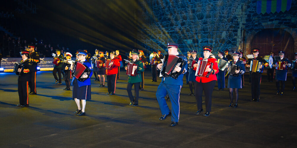 Group of uniformed musicians playing accordions in a night-time outdoor performance. They are standing in formation in 5 rows and in front of a large grey castle backdrop.