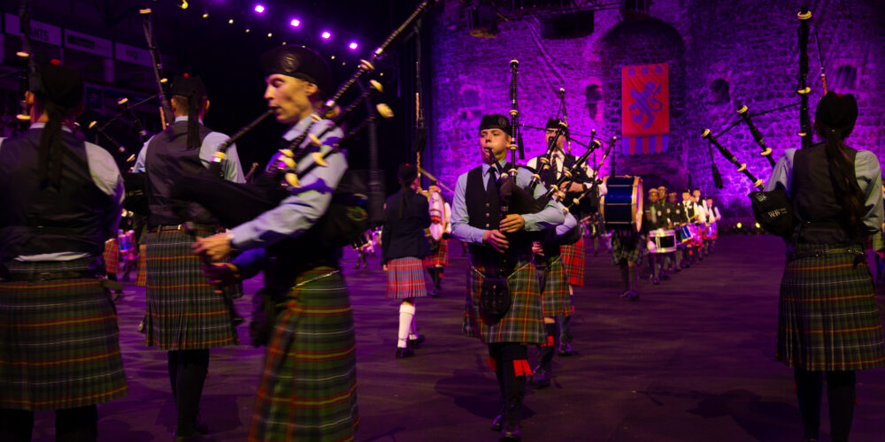 A large group of bagpipers in traditional Scottish kilts and attire stand in various rows, performing under purple stage lights at an outdoor event with a castle backdrop.