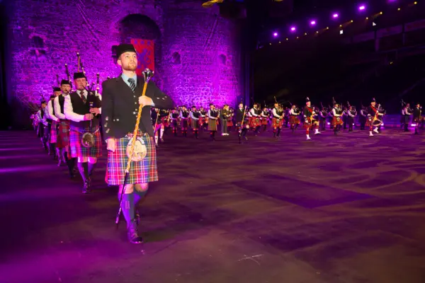 A large group of bagpipers and drum majors in traditional Scottish kilts and accompanying attire. They are performing indoors under purple stage lighting with a castle backdrop behind the musicians.