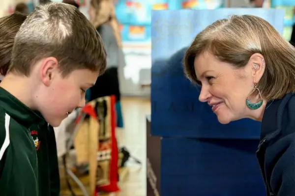 A young boy in a dark green jacket and a woman in a navy fleece smiling warmly at each other in a brightly lit indoor setting.