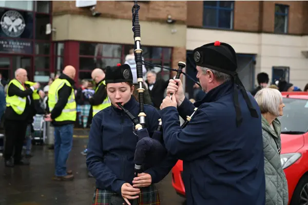 Two bagpipers in traditional attire playing outdoors during a public event with onlookers in the background.