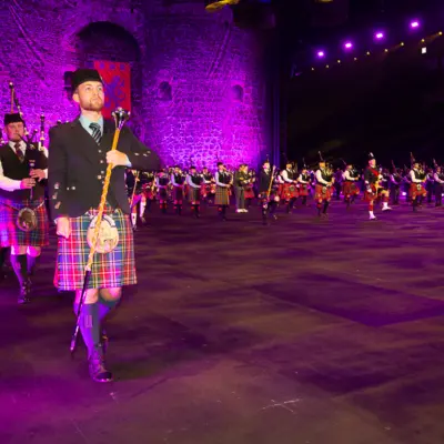 A large group of bagpipers and drum majors in traditional Scottish kilts and accompanying attire. They are performing indoors under purple stage lighting with a castle backdrop behind the musicians.