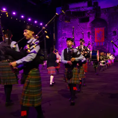 A large group of bagpipers in traditional Scottish kilts and attire stand in various rows, performing under purple stage lights at an outdoor event with a castle backdrop.