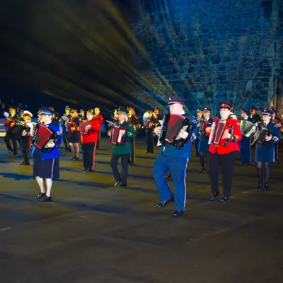 Group of uniformed musicians playing accordions in a night-time outdoor performance. They are standing in formation in 5 rows and in front of a large grey castle backdrop.