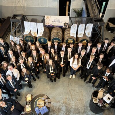 A large group of students and adults gathered in a brewery setting with barrels and banners overhead.