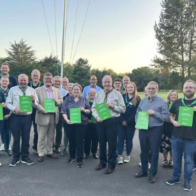 Group of smiling adults standing outdoors holding green workbooks in a park-like setting during daylight.