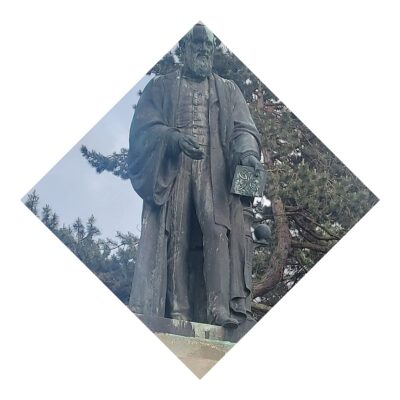 Statue of a robed figure, Lord Kelvin, holding a book with intricate designs. The statue is standing outdoors against a backdrop of pine trees and a cloudy sky.